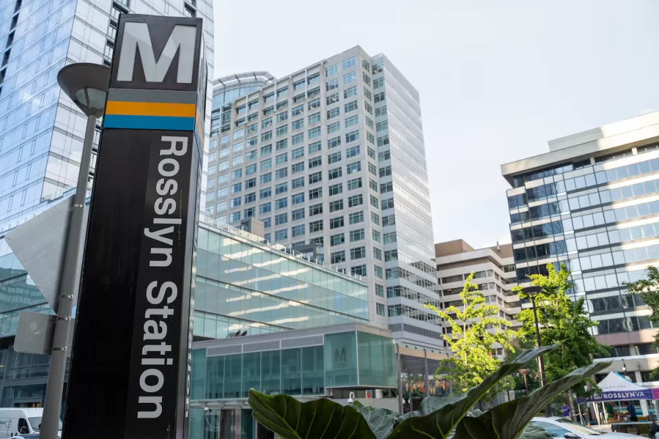 Close-up of Metro sign column at Rosslyn Station in Alexandria, Virginia with high-rose glass buildings in background.